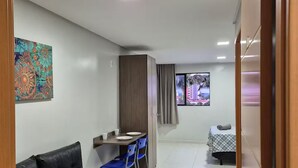 Un salon moderne avec un canapé en cuir noir, une table à manger avec des chaises bleues, un petit coin cuisine et une chambre avec un lit et des rideaux.
