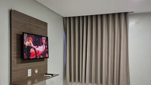 Une chambre d’hôtel moderne dotée d’un téléviseur à écran plat fixé sur un panneau en bois, d’un lit avec une couette à carreaux et d’une fenêtre avec des stores verticaux.