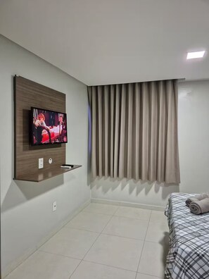 Ein modernes Hotelzimmer mit einem an einer Holzplatte montierten Flachbildfernseher, einem Bett mit einer Karo-Bettdecke und einem Fenster mit vertikalen Jalousien.