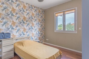 Ein Schlafzimmer mit einem Bett, einer Kommode, einem Fenster mit Jalousien und einer Wand mit einem tropischen Blattmuster.