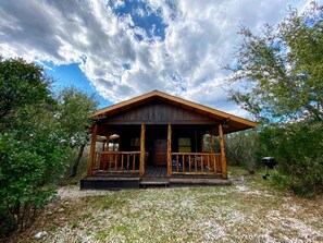 A rustic cabin with a porch, surrounded by trees and a clear sky. - Side by Side Cabins on a Texas Ranch, Perfect for a Family Glamping Getaway from San Antonio (Mico)