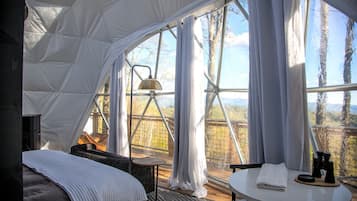 A tented accommodation with a bed, a small table with a lamp, and a view of the outdoors.