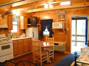 Interior de una cabaña con paredes de madera, una cocina con estufa, refrigerador y una mesa de comedor con sillas.