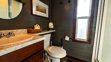 A bathroom with a marble sink, wooden cabinets, a toilet, a framed picture on the wall, and a window.