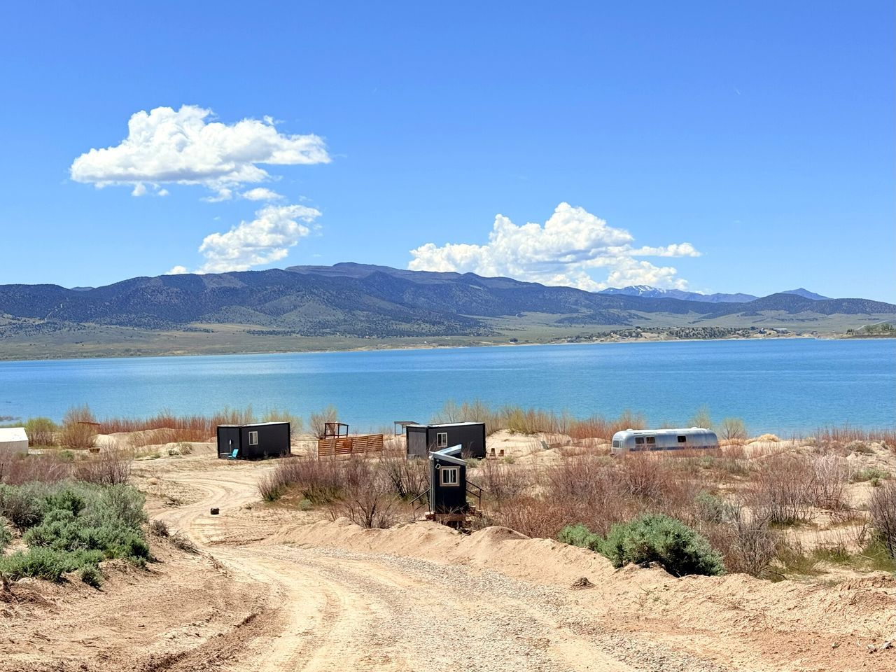 A campsite with a dirt path leading to a small cabin and a trailer by a lake with mountains in the background.