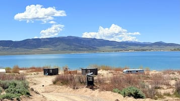A campsite with a dirt path leading to a small cabin and a trailer by a lake with mountains in the background.