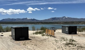 Paisaje desértico con dos cabañas negras, un banco de madera y una cadena montañosa al fondo.