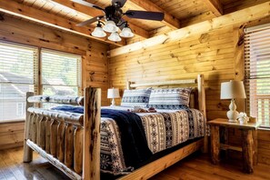 Ein Schlafzimmer einer Holzhütte mit einem Bett, zwei Nachttischen, einem Deckenventilator und großen Fenstern.