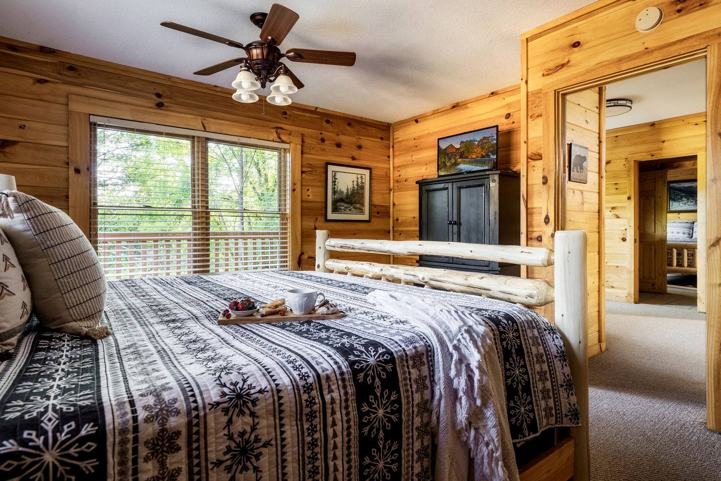 Een slaapkamer in een houten hut met een bed, een plafondventilator, een raam, een televisie en een deur die naar een andere kamer leidt.