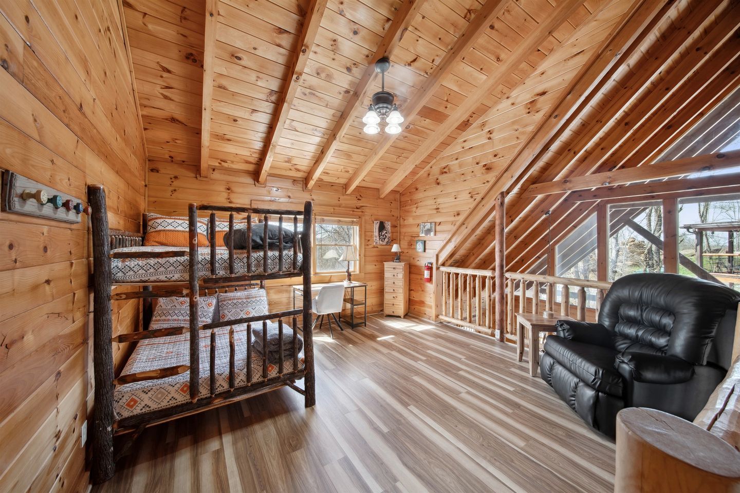 Interior de una cabaña de madera con literas, un escritorio, una silla y vistas al exterior.