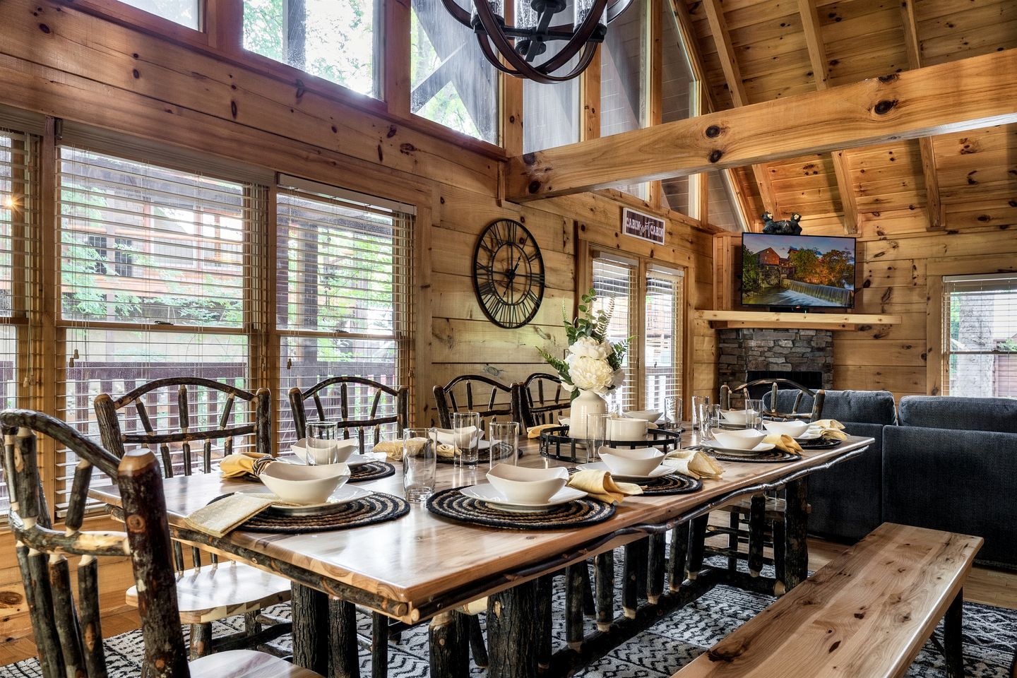A wooden dining room with a large table set for six, surrounded by chairs, and a stone fireplace.