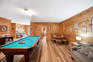 Een pooltafel, een eettafel met stoelen, een bank en houten wanden met verschillende borden.