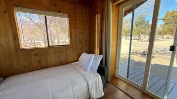 A single bed in a wooden cabin room with a large window and a sliding glass door leading to an outdoor area.