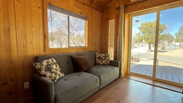 Interior de una cabaña de madera con sofá, cojines y un ventanal grande.