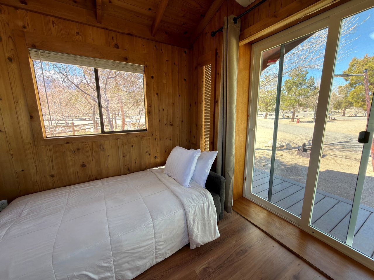 A single bed in a wooden cabin room with a large window and a sliding glass door leading to an outdoor area.