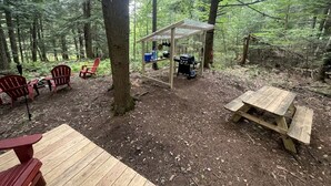 A forested campsite with a wooden table, red Adirondack chairs, a covered picnic table, and a grill.