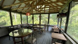 A covered outdoor dining area with wooden tables and chairs, large windows, and a view of trees.