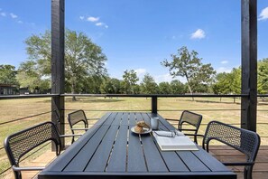 Una mesa de madera al aire libre con sillas, un perro y un libro en una terraza con vistas a un campo de césped y árboles.