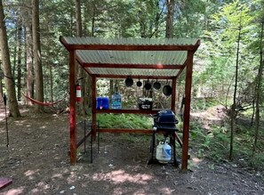 A rustic wooden structure with a corrugated metal roof, a table with hanging pots, and a water container.