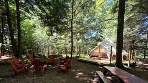 Un terrain de camping aménagé dans une forêt, avec une tente, des chaises Adirondack rouges et une table de pique-nique.