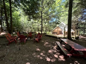 A forest campsite with a tent, red Adirondack chairs, and a picnic table.