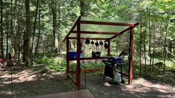 Una estructura de madera con techo metálico, un banco y un colgador para ollas en un área boscosa.