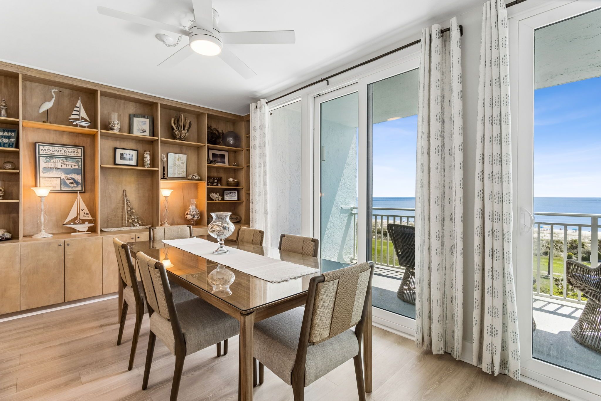 Un comedor con una mesa y sillas de madera, un ventilador de techo y vistas al océano a través de amplios ventanales correderas.