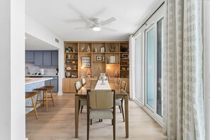 A dining area with a wooden table and chairs, a kitchen in the background, and a large window with curtains. - 1372 Shipwatch Oceanfront Beautifully Updated (Fernandina Beach)