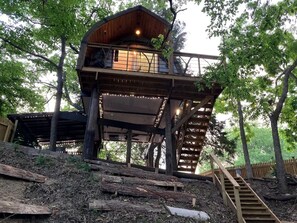 Ein Baumhaus aus Holz mit einem Balkon, umgeben von Bäumen und einer Holztreppe.