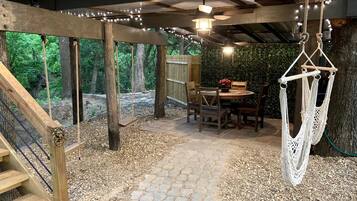 A covered outdoor area with a stone pathway, wooden deck, hanging macrame chair, and a table with chairs.