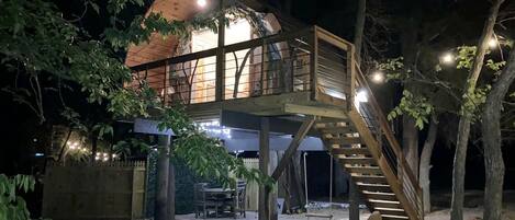 A wooden house with a staircase leading to a deck, surrounded by trees and a fence.