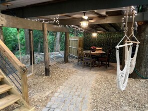 Un espacio al aire libre cubierto con un sendero de piedra, terraza de madera, silla colgante de macramé y una mesa con sillas.