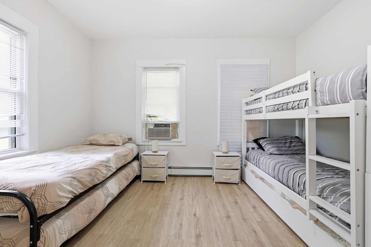 A bedroom with a single bed, a bunk bed, a bedside table with drawers, an air conditioning unit, and a window with blinds.