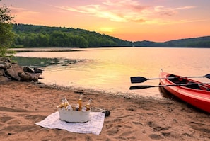 Escena junto al lago con un kayak y un balde de bebidas embotelladas sobre la arena.