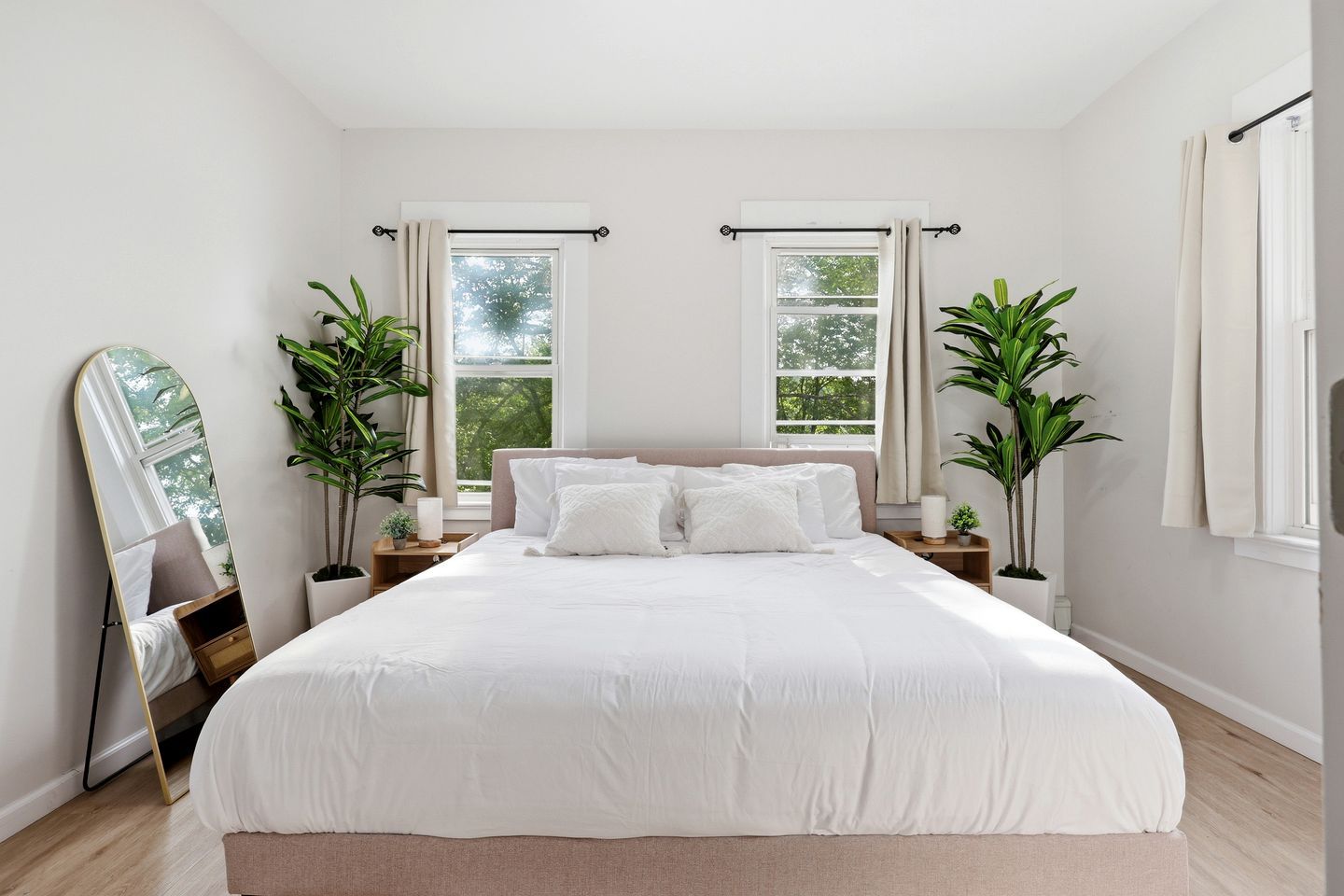 A bedroom with a large bed, two windows with curtains, a bedside table with a lamp, and two potted plants.