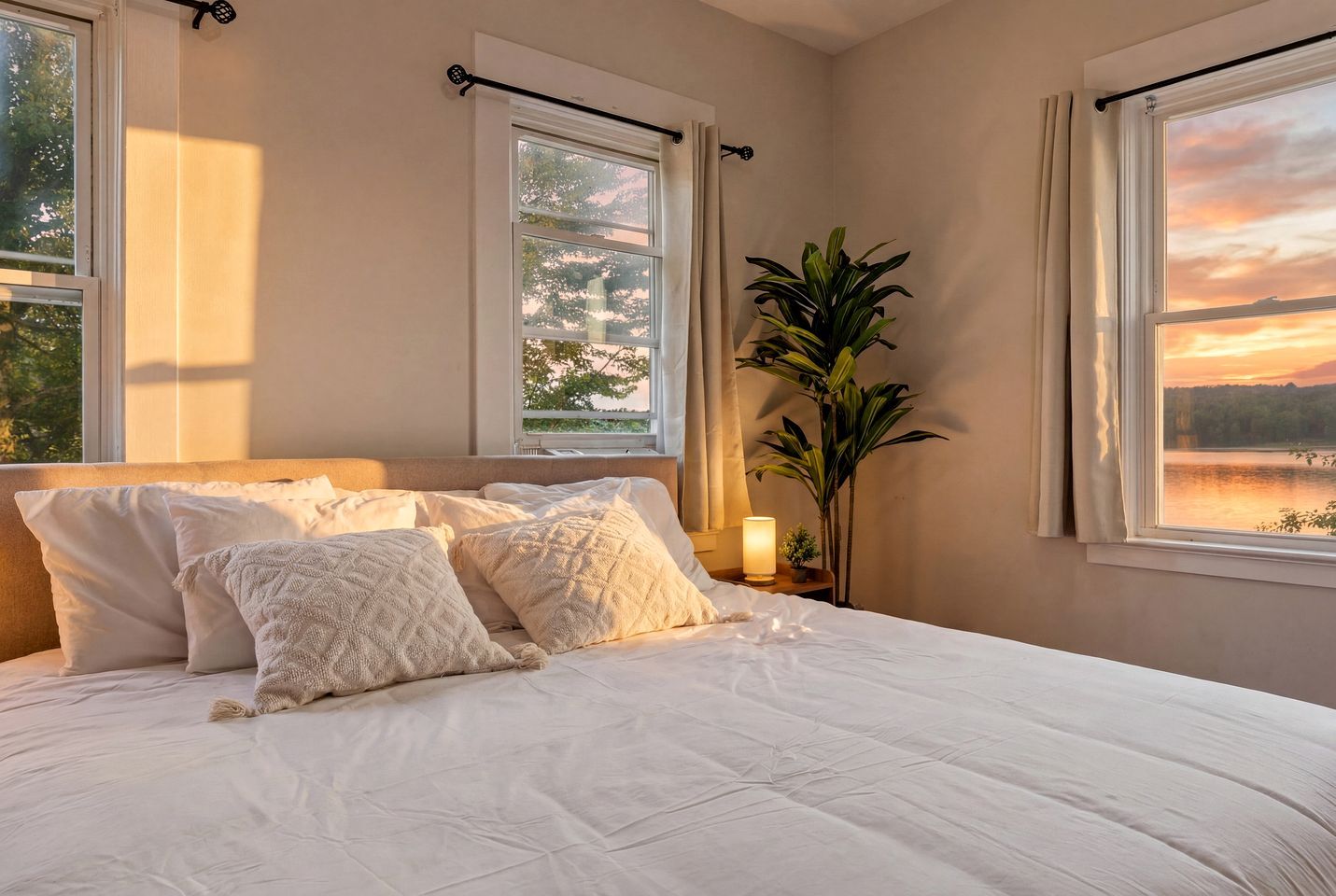 A bedroom with a large bed, two windows, a nightstand with a lamp, and a potted plant.