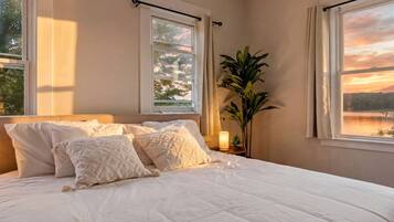 A bedroom with a large bed, two windows, a nightstand with a lamp, and a potted plant.
