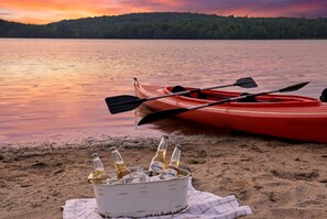 Un kayak y botellas sobre una playa arenosa con un lago y colinas al fondo.