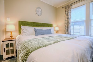 A neatly made bed with a green headboard, two bedside lamps, a small cabinet, and a window with curtains.