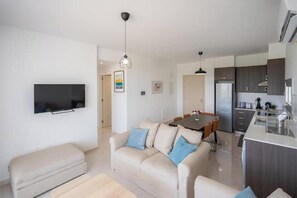 Ein modernes Wohnzimmer mit Flachbildfernseher, einer Couch, einem Esstisch und einer Küchenzeile inklusive Kühlschrank.