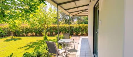 En overdækket terrasse med bord og stole, en dør og en velplejet græsplæne.