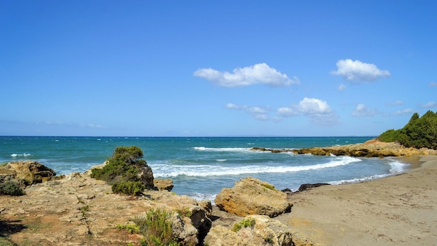 Paisagem costeira com terreno rochoso, uma praia de areia e um céu azul límpido.