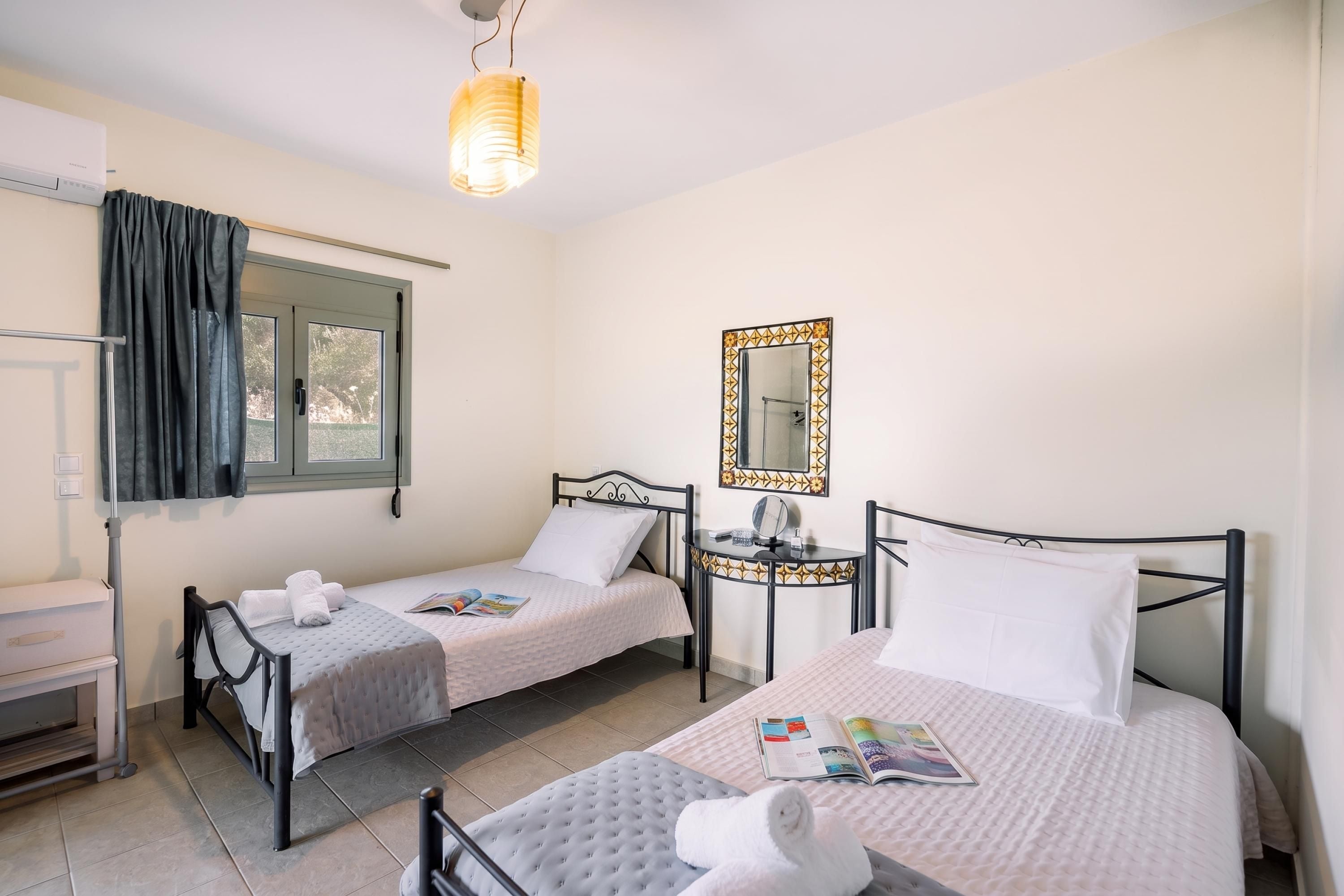Hotellihuone, jossa on kaksi erillistä sänkyä, pieni pöytä peilillä, ikkuna verhoilla ja kattovalaisin.