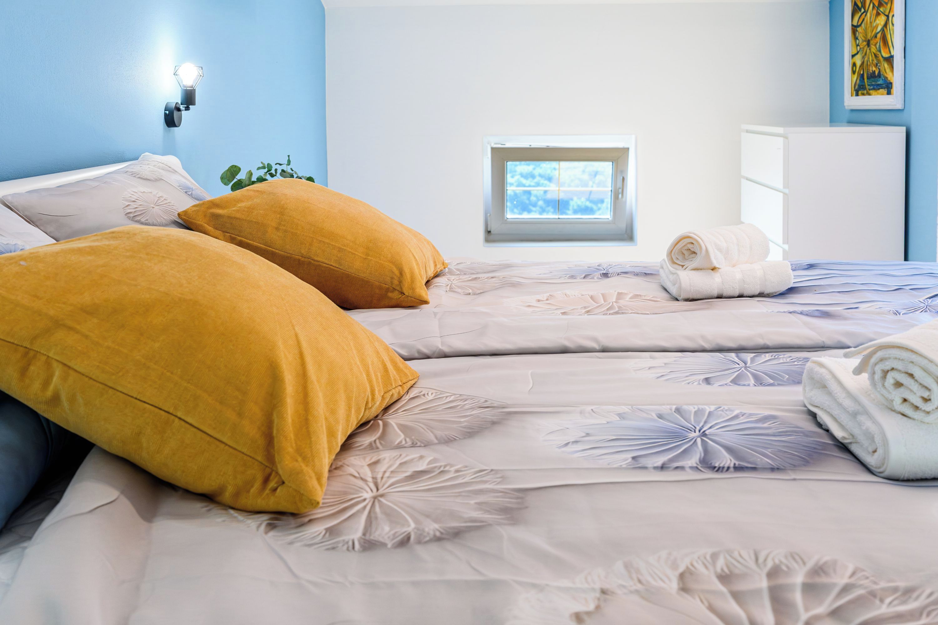 Un dormitorio con una cama, dos almohadas, una ventana, una toalla y una lámpara fijada a la pared.