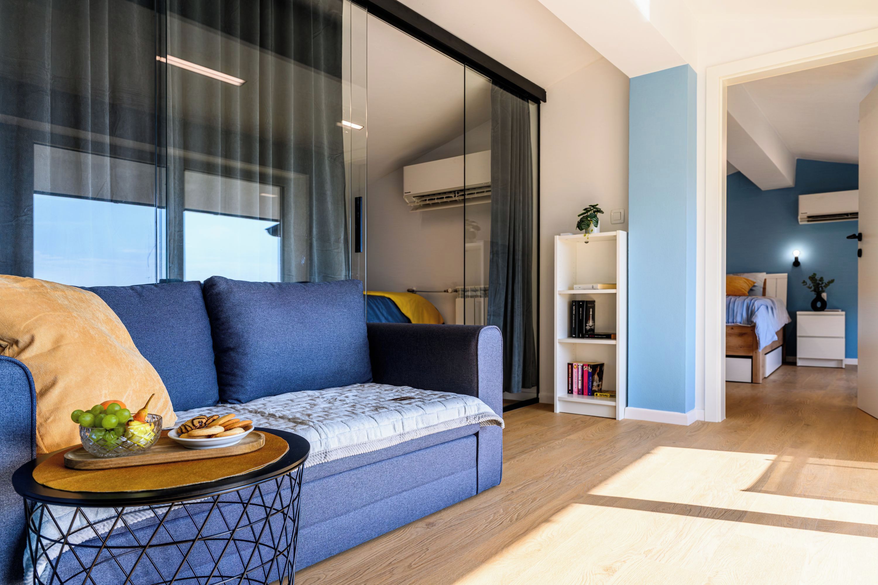 A modern living room with a sofa, a side table with a fruit bowl, a bookshelf, and a view into another room with a bed and a nightstand.