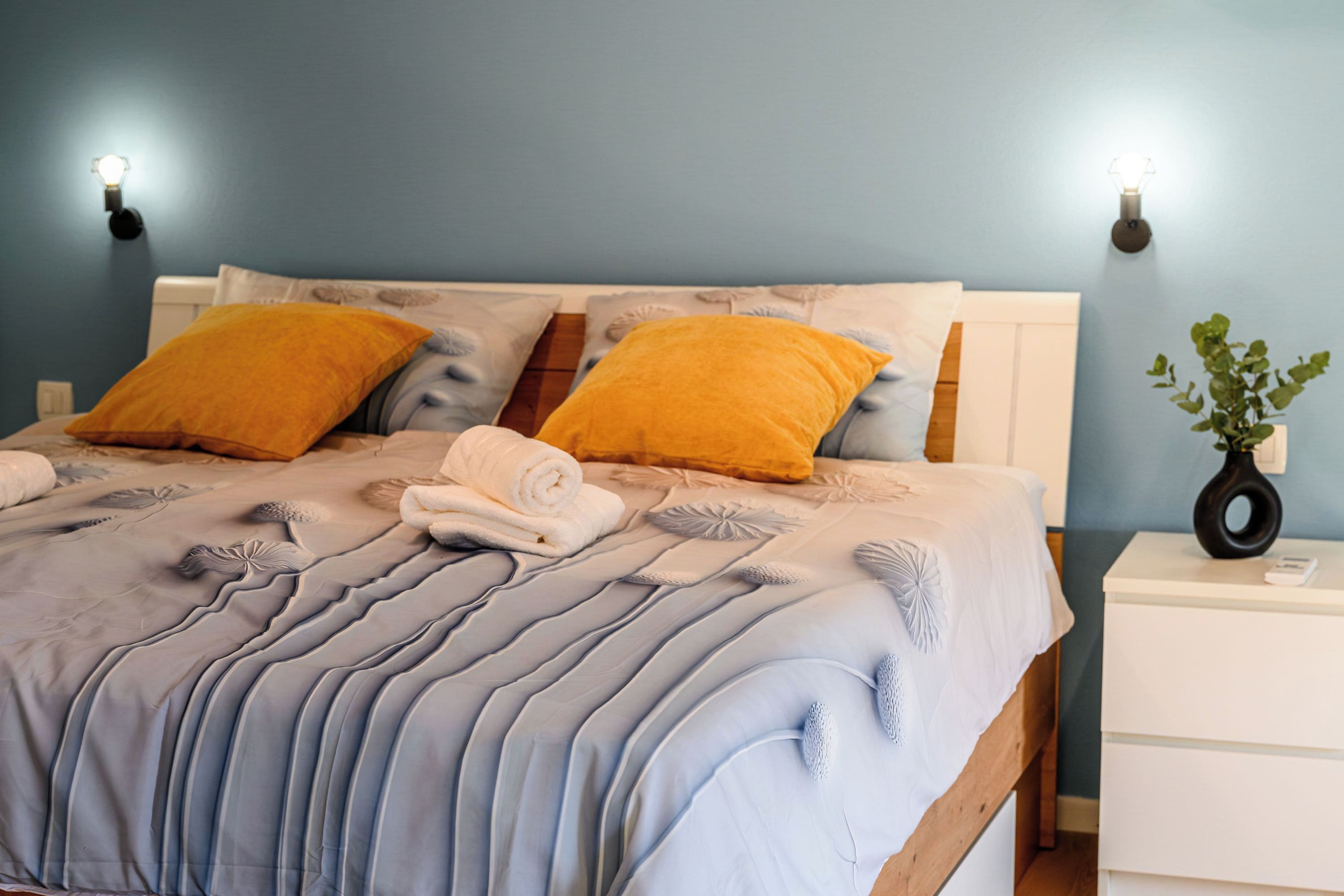 Ein Bett mit einer blau-weißen Tagesdecke, zwei gelben Kissen und einem weißen Nachttisch mit einer Pflanze.