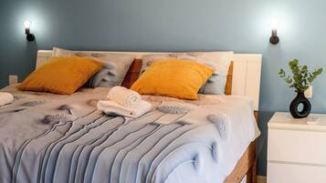 Ein Bett mit einer blau-weißen Tagesdecke, zwei gelben Kissen und einem weißen Nachttisch mit einer Pflanze.