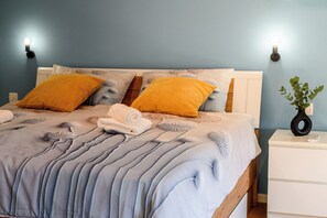 Een bed met een blauw-witte spreidek, twee gele kussens en een witte nachtkastje met een plant.