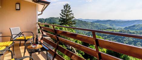 A balcony with a wooden railing, yellow cushioned chairs, a table with a bowl and wine glass, and a view of a mountainous landscape.