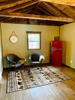 Une pièce avec des poutres en bois au plafond, deux chaises, un réfrigérateur rouge, une horloge murale et un tapis à motifs.